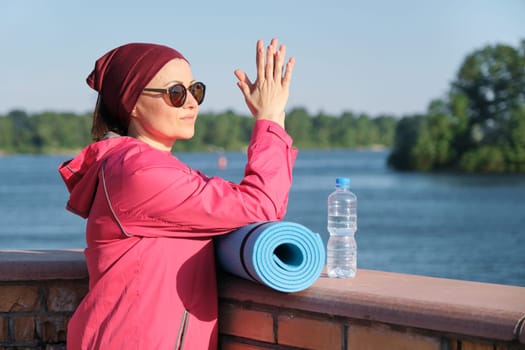 Healthy lifestyle of mature woman, outdoor portrait of an age female in sportswear with yoga mat and bottle of water, background blue sky, river, evening sun