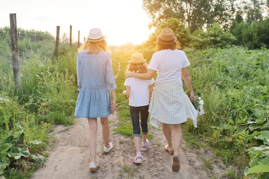 Mother with children two daughters walking along a country road, background summer meadow sunset, back view