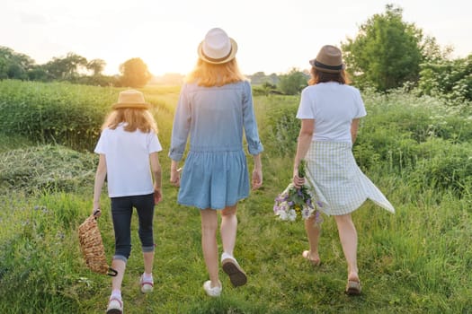 Mother with children two daughters walking along a country road, background summer meadow sunset, back view