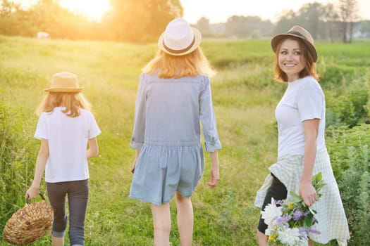 Mother with children two daughters walking along a country road, background summer meadow sunset, back view