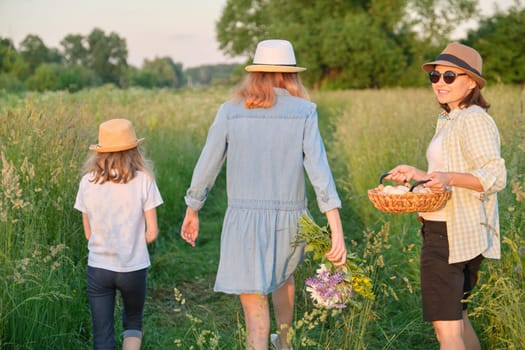 Woman with two daughters walking in a meadow with a basket of eggs, back view, golden hour.