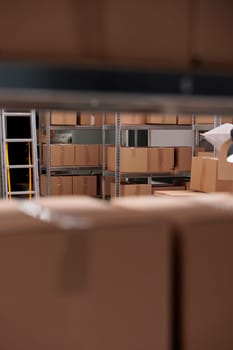 Selective focus of tall metal shelves filled with various cardboard boxes and containers, empty industrial warehouse. Storehouse building used for storage and distribution centers