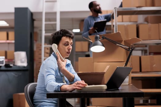 Caucasian manager talking at landline phone with remote supervisor discussing transportation logistics problem while looking at cargo stock on laptop. Employee working at order delivery in warehouse