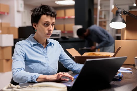 Storehouse manager sitting at desk table looking at online orders before start preparing customers packages, checking cargo stock. Supervisor woman working in warehouse delivery department