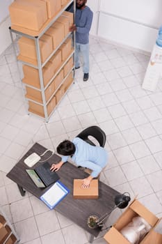 Top view of storehouse manager standing at desk in warehouse while preparing customer order putting clothes in cardboard box after checking delivery detalies. Team working in distribution center
