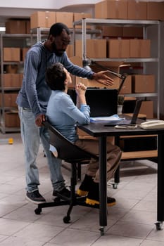 Workers analyzing transportation logistics while checking metallic box delivery order on laptop before shipping package. Multi ethnic team working in warehouse, looking at cargo stock