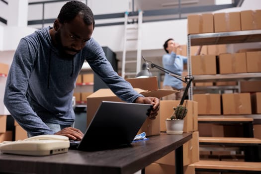 African american supervisor looking at cargo stock on laptop computer before start preparing customers orders, working in warehouse delivery department. Worker checking shipping detalies