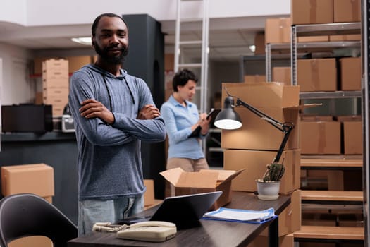 Portrait of african american employee standing with arm crossed in warehouse while working at customers packages delivery, preparing carton boxes. Distribution center fulfillment company