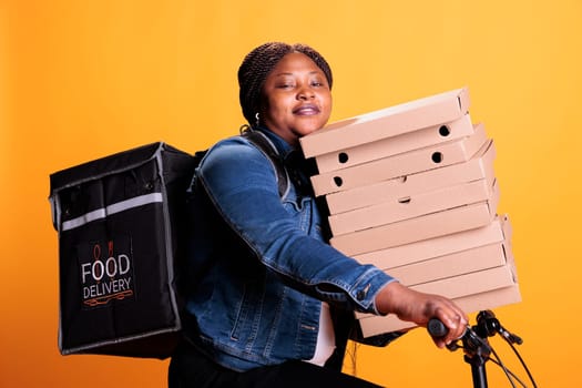 Pizzeria delivery worker carrying stack of cardboard full with pizza delivering takeaway food order to customer during lunch time, standing in studio. Restaurant employee using bike as transportation