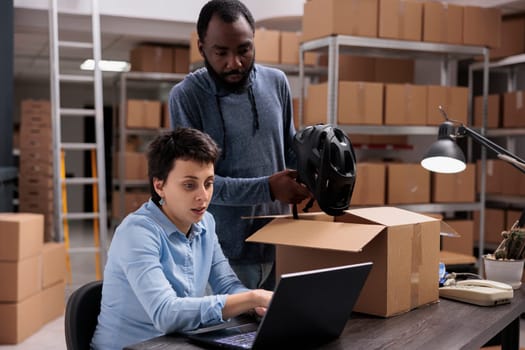 Storehouse employee putting customer helmet order in carton box preparing delivery for client while discussing transportation details with manager. Diverse team working in warehouse