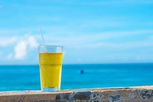 Big glass drink plastic straw green tea looks like yellow beer or kombucha blue sea white clouds background water drops condensed.