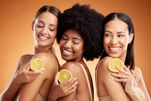 Skincare, diversity and women, beauty and lemon for health, wellness and nutrition on orange studio background. Friends, smile and happy models with fruit for vitamin c, healthy or glowing skin