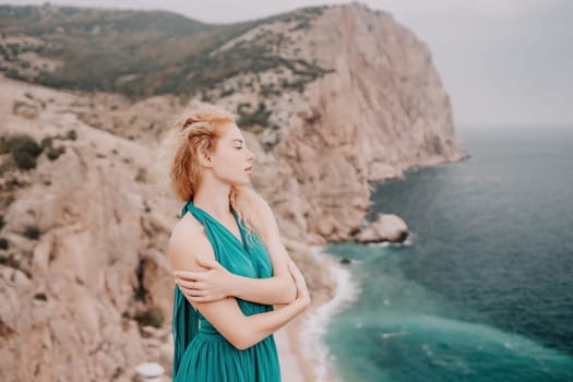 Side view a Young beautiful sensual woman in a mint long dress posing on a volcanic rock high above the sea during sunset. Girl on the nature on overcast sky background. Fashion photo