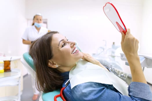 Process of dental treatment. Female patient looking at her teeth in mirror while sitting in dental chair. Healthcare, medical and dentistry concept