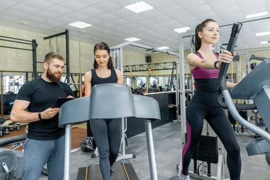 Young smiling fitness women with personal trainer an adult athletic man on treadmill in the gym. Sport, teamwork, training, healthy lifestyle concept