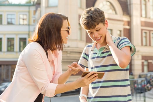 Parent and teenager, relationship. Mother shows her son something in mobile phone, boy is embarrassed, smiling, holding his hands on his head, city street background.