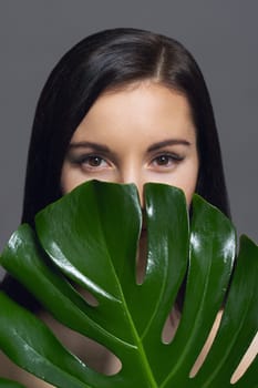 Studio beauty portrait of young brunette with natural make-up perfect skin with green exotic leaf on gray background. Concept of natural cosmetics, face and body care products