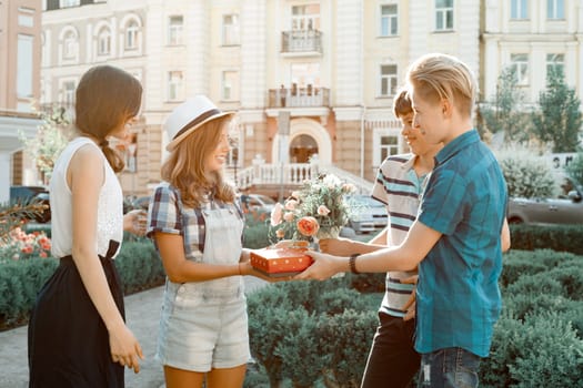 Outdoor portrait of happy group of teenagers friends congratulating girl with bouquet of flowers and gift in box. Friendship and people concept.