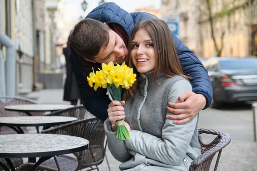 Couple in love in city. Young happy man and woman in spring city talking, walking, with bouquet of yellow flowers daffodils.