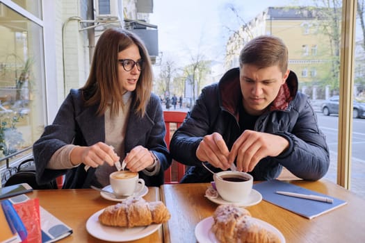 Young male and female friends students sitting in outdoor cafe, talking, drinking coffee, tea, eating croissants. On table textbooks, notebooks, city background