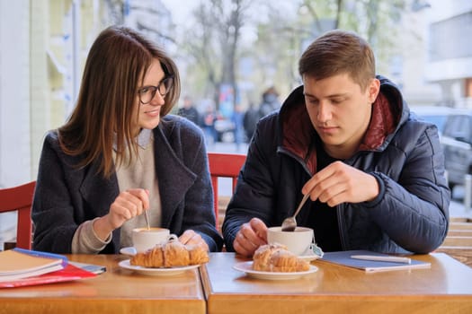 Young male and female friends students sitting in outdoor cafe, talking, drinking coffee, tea, eating croissants. On table textbooks, notebooks, city background