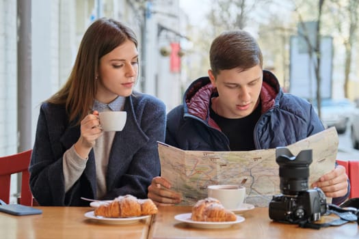 Young tourists man and woman reading map of city in outdoor cafe. Couple drinking coffee tea and eating croissants, spring city background.