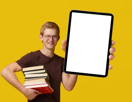 Young student holding a tablet PC with a white blank screen, surrounded by books, studying and using modern technology for academic purposes. Blond man smiles with PDA isolated on yellow background. High quality photo