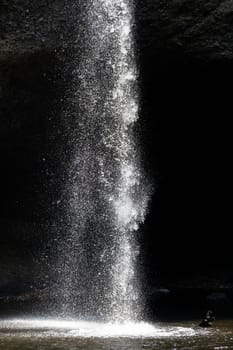 Splashing waterfall drop on pond with dark background of cave in summer.