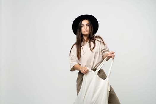 Brunette woman in a beige t-shirt and black hat with a cotton shopper bag with groceries looking in a camera. Reusable eco bag for shopping. Zero waste concept