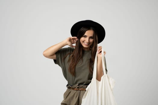 Happy smiling brunette woman in black hat with white cotton eco bag on her shoulder. Girl holding textile grocery bag with vegetables. Zero waste concept