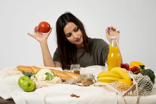 Happy woman in green t-shirt unpacking shopping mesh eco bag with healthy vegetables, fruits, bread, snacks on the kitchen at home. Healthy eating vegetarian concept