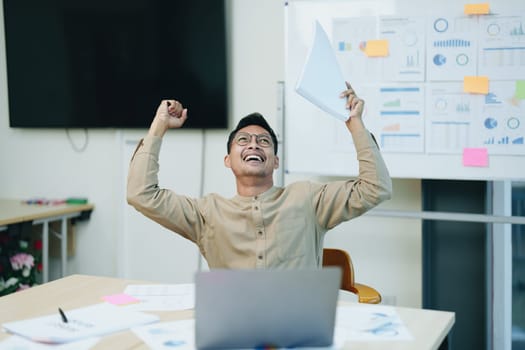 Portrait of a male business owner showing a happy smiling face as he has successfully invested his business using computers and financial budget documents at work.