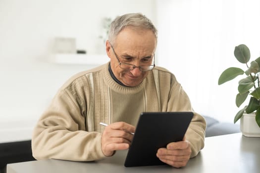 Senior man reading news on digital tablet. Serious mature male using portable computer at home, sitting at table, copy space