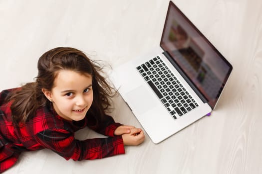 Top view photo of beautiful little girl lying on wooden floor. Girl looking at camera and using laptop