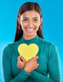 Portrait, makeup and heart on valentines day with a woman in studio on a blue background for love or health. Face, beauty and romance with an attractive young female holding a cut out emoji or symbol.