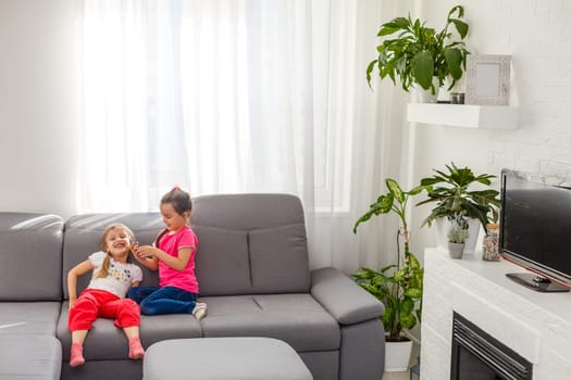 Stay Home Stay Safe. Two happy kids sitting on cozy chair relaxing playing in white living room indoors. Sisters having fun at home