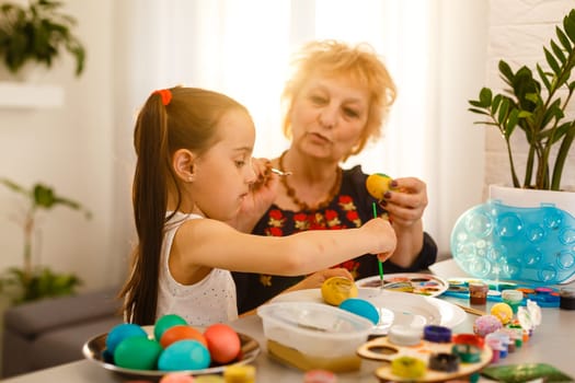 Little girl and her grandmother painting Easter eggs at home