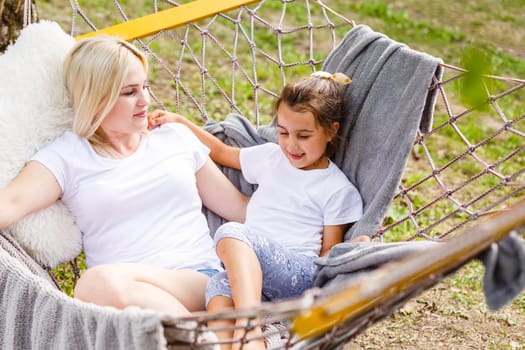 Wide view of a young mother and daughter relaxing together and smiling sitting in a hammock, hugging and lounging during a sunny summer day in a holiday home garden with grass and trees, lifestyle.