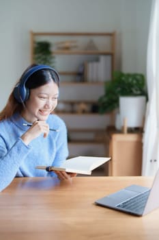 Portrait of a teenage Asian woman using a computer, wearing headphones and using a notebook to study online via video conferencing on a wooden desk at home.