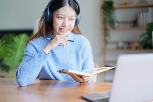 Portrait of a teenage Asian woman using a computer, wearing headphones and using a notebook to study online via video conferencing on a wooden desk at home.