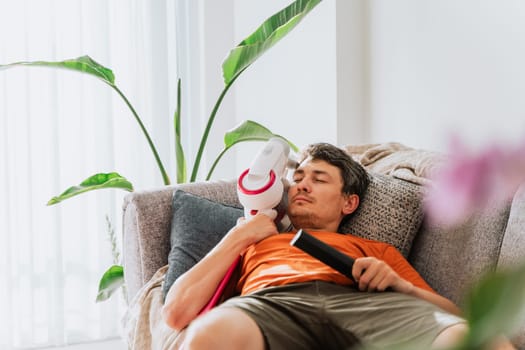 Tired man sleeping on a couch after cleaning the house holding TV remote control and wireless vacuum cleaner. White caucasian male relaxing taking nap on a sofa watching TV with plants in background.