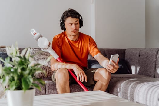 Man with wireless vacuum cleaner and headphones sitting on sofa checking messages in cellphone. White caucasian male tired of cleaning texting in smartphone holding vacuum cleaner wearing earphones.