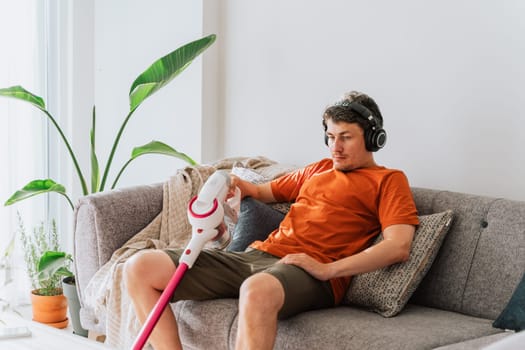 Tired man with wireless vacuum cleaner relaxing chilling on the couch surrounded with plants. White caucasian male exhausted after spring cleaning sitting on sofa white light room plants in the back.