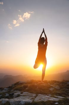 Outdoor yoga silhouette, young woman in tree pose. A woman practices yoga on the green grass on top of a mountain with a beautiful sunset or sunrise view. Tree pose or vrikshasana with arms raised together above the head. Rest, harmony with nature.