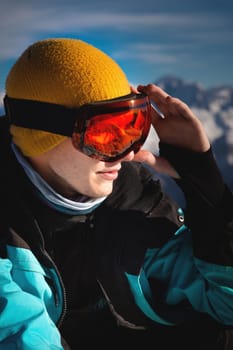 A young guy skier in ski goggles with the reflection of the sun in them, stands in the mountains and looks away holding his hand to his head, portrait.