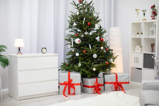 A bright living room with live Christmas tree decorated in white and red. Silver gifts with red ribbons.