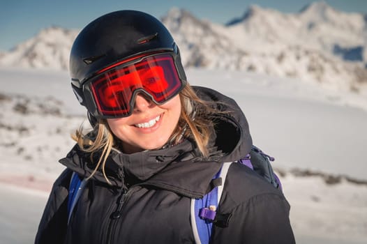 A skier smiles in a safety ski helmet and goggles against the backdrop of the picturesque Alpine mountains. Active sports people and success concept image.