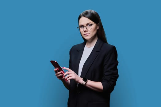 Young business woman using smartphone on blue color studio background. Serious confident female in glasses jacket looking at camera. Mobile applications, digital technologies in business work leisure