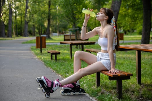 Spending time in a green city park. A young woman sits on a bench and drinks from a bottle. A hot day in nature. Athlete rests during training while sitting on a bench