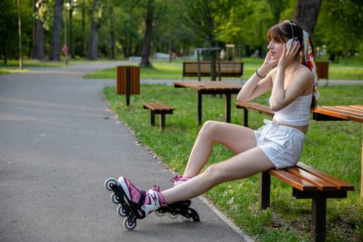 Skater girl resting on a park bench. A girl wearing rollerblades. A young woman with wireless headphones on her head. Trees and lawns and an asphalt alley are visible in the background.